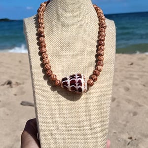 May include: A brown beaded necklace with a large, patterned shell pendant. The necklace is displayed on a beige bust against a backdrop of a beach with blue water and sky. The beads appear to be approximately 0.5 inches in diameter.