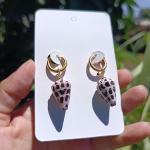 Puede incluir: Un par de pendientes de aro dorados con conchas marinas en forma de cono que cuelgan, con un patrón natural de manchas marrones y crema. Los pendientes están sujetos a una tarjeta de presentación rectangular blanca de color dorado.
