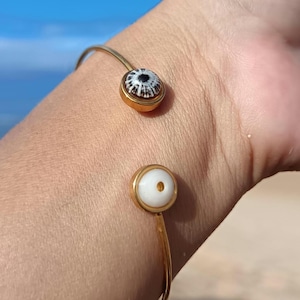Può includere: Un bracciale aperto color oro con due elementi decorativi circolari. Uno ha un motivo in bianco e nero, l'altro è bianco con un foro centrale. Il bracciale è indossato su un polso.