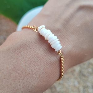 Peut inclure: Un bracelet doré à perles avec un motif central de coquillages blancs empilés. Le bracelet est délicat et semble fait à la main, mettant l'accent sur les éléments naturels et une esthétique de plage.
