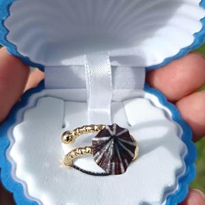 May include: A gold-colored ring with a seashell design, presented in a blue seashell-shaped box. The ring features a textured band and a dark brown and white seashell accent, perfect for ocean-themed jewelry.