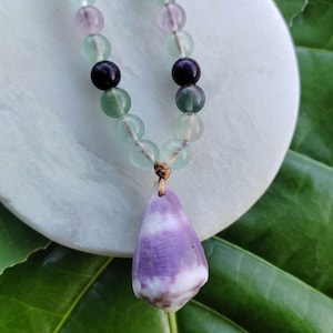 Puede incluir: Un collar artesanal con un colgante y una cadena de cuentas. El colgante tiene forma de lágrima, con tonos morados y blancos. La cadena de cuentas alterna entre cuentas verdes, moradas y negras. El collar se exhibe sobre una superficie circular blanca.