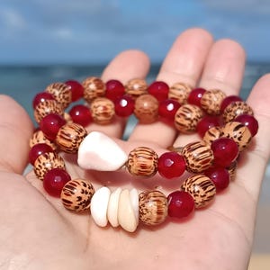 Peut inclure: Deux bracelets de perles sur le thème de la plage. L'un comporte des perles rouges facettées et des perles en bois à motifs. L'autre a des perles similaires, ainsi que des accents de coquillages blancs. Les bracelets sont tenus dans une main.
