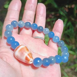 May include: A blue beaded bracelet with a seashell pendant. The bracelet features round, light and dark blue beads. The pendant is a white and orange seashell. The bracelet is held in a hand.