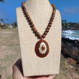 May include: A handcrafted necklace featuring a pendant made from a brown shell with a white and orange center, suspended from a strand of wooden beads. The necklace is displayed on a beige mannequin against a backdrop of a sunny beach.