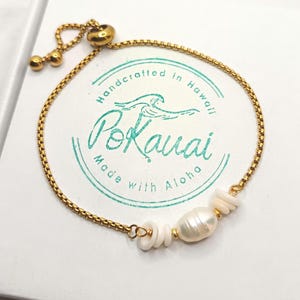 May include: Gold-toned chain bracelet with a central pearl and white shell accents. The bracelet features an adjustable clasp and is presented on a white surface. The bracelet is handcrafted in Hawaii.