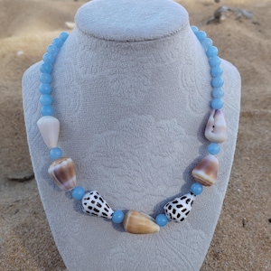Puede incluir: Collar con cuentas azul claro y diversas conchas marinas en blanco, marrón y negro. El collar se exhibe en un maniquí gris claro. El diseño evoca un tema natural y costero, ideal para el verano.
