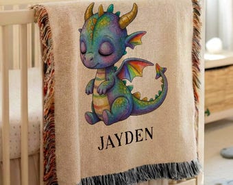 May include: A woven throw blanket featuring a colourful illustration of a baby dragon with purple, green, and orange scales. The name JAYDEN is printed in black capital letters below the dragon. The blanket has fringed edges with multi-coloured threads.