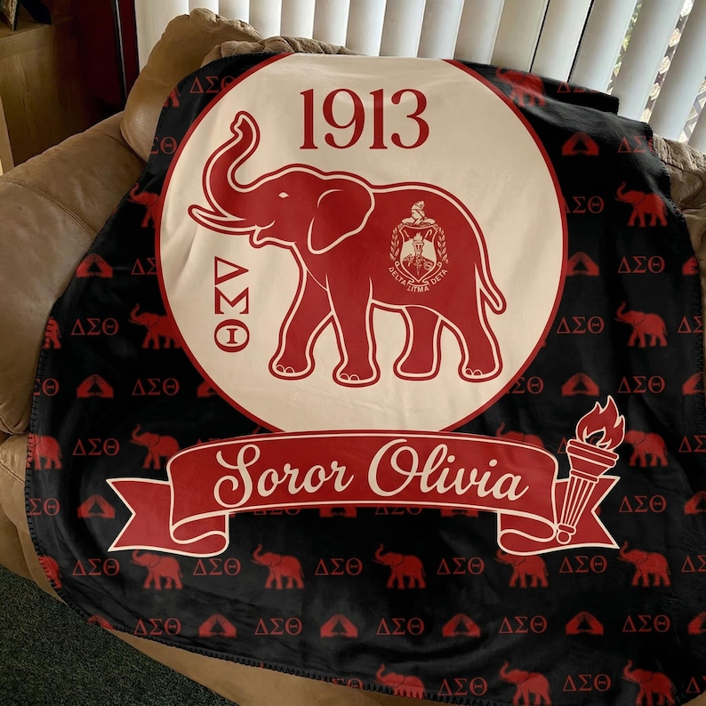 Puede incluir: Una manta negra con un gran emblema circular de color crema que presenta un elefante rojo, el n&uacute;mero 1913 y letras griegas. Una cinta roja en la parte inferior muestra el texto Soror Olivia, junto a una antorcha y patrones de elefantes.