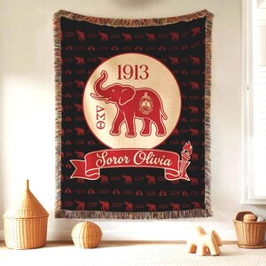 Puede incluir: Una manta tejida negra con un patr&oacute;n repetitivo de elefantes rojos y letras griegas. Un gran c&iacute;rculo central muestra el a&ntilde;o 1913, un elefante rojo y el texto Soror Olivia sobre una cinta roja con un icono de antorcha en el extremo.
