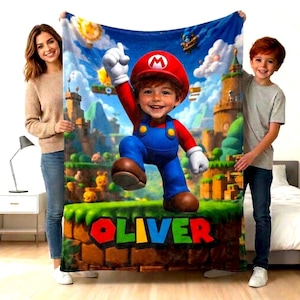 Puede incluir: Una manta rectangular de forro polar con un gráfico vibrante y colorido de un niño disfrazado de Mario de la serie de videojuegos Super Mario, sobre un paisaje fantástico con el nombre OLIVER impreso en letras mayúsculas multicolores.