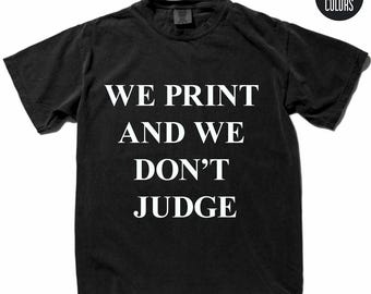 May include: A black short-sleeved t-shirt featuring the white text WE PRINT AND WE DON'T JUDGE centred on the chest. A circular black logo with the white text COMFORT COLORS is positioned in the upper right corner of the image.