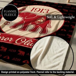 Puede incluir: Una manta de forro polar de franela en negro y crema con un gr&aacute;fico grande de un elefante rojo, el a&ntilde;o 1913 y un emblema circular. El patr&oacute;n de fondo incluye siluetas de elefantes rojos y letras griegas. El reverso es de color crema liso.