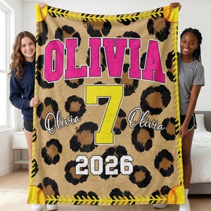 Puede incluir: Una manta rectangular de forro polar con un fondo de estampado de leopardo en beige y negro. El nombre OLIVIA está impreso en grandes letras rosas de estilo universitario, un gran número 7 amarillo está centrado y el año 2026 aparece en blanco. Costuras de pelota de sóftbol amarillas bordean los lados.