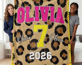 May include: A rectangular fleece blanket featuring a tan and black leopard print background. The name OLIVIA is printed in large pink varsity letters, a large yellow number 7 is centred, and the year 2026 is in white. Yellow softball stitching borders the edges.