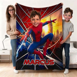 Puede incluir: Una manta rectangular de forro polar con el gráfico de un niño disfrazado de Spider-Man en una pose dinámica sobre un fondo de horizonte urbano y un patrón de telaraña. El nombre MARCUS está impreso en letras rojas.