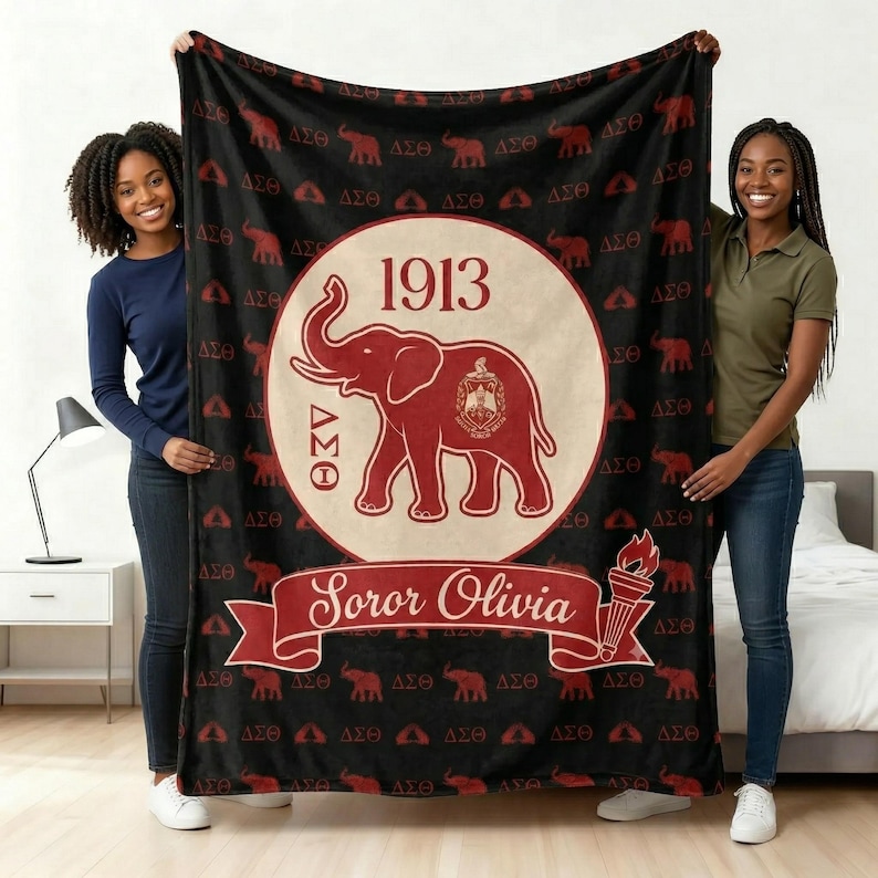 Puede incluir: Una manta de forro polar negra con un patr&oacute;n repetitivo de elefantes rojos y las letras griegas Delta Sigma Theta. Un gran c&iacute;rculo central muestra un elefante rojo, el a&ntilde;o 1913 y una pancarta con el nombre Soror Olivia y una antorcha.