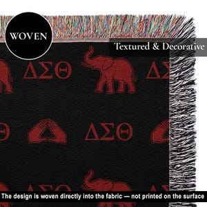 Puede incluir: Una manta tejida de color negro con un patr&oacute;n repetitivo de elefantes rojos, las letras griegas Delta Sigma Theta y s&iacute;mbolos de manos. La manta tiene una superficie texturizada con flecos multicolores en los bordes superior y lateral.