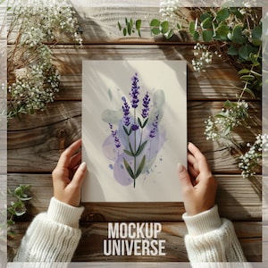 May include: A watercolor painting of lavender flowers on a white background. The painting is framed and held by a person's hands. The background is a wooden surface with green plants.