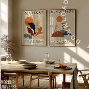 May include: Two framed Matisse art prints with gold frames hang above a wooden dining table. One print features abstract shapes in orange, blue, and red. The other depicts dancers. A vase, plates, and fruit are on the table.