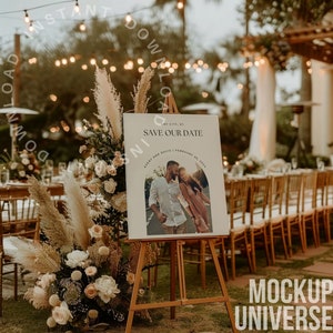 May include: A white sign with the text "Save Our Date" and a photo of a couple in front of a blurred background of a wedding reception with white chairs and a greenery archway. The sign is on a wooden easel.