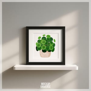 Può includere: Una stampa artistica incorniciata con un'illustrazione ad acquerello di una pianta di Pilea peperomioides, nota anche come pianta dei soldi cinese. L'opera d'arte mostra una pianta verde vibrante in un vaso beige, su sfondo bianco, incorniciata in una cornice quadrata nera.