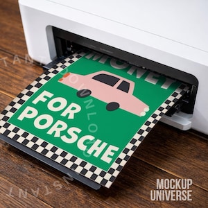 Peut inclure: Une imprimante blanche imprime une affiche verte avec un dessin de voiture rose et les mots "MONEY FOR PORSCHE". L'affiche a une bordure à carreaux noirs et blancs et est sur une surface en bois.