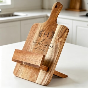 May include: A wooden cutting board with a handle and a built-in stand. The board is engraved with the text "H The Howards est 2020" and a decorative floral design. The board is made of light and dark brown wood.