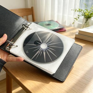 May include: A black textured binder with metal rings holding clear plastic sleeves. Inside the sleeves are circular discs, including one black disc featuring a prominent white starburst or sunburst graphic design radiating from the center hole.