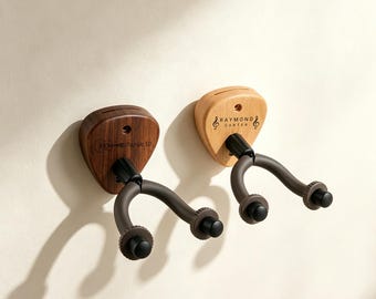 May include: Two wall-mounted guitar hangers with pick-shaped wooden bases. The left base is dark wood with a guitar icon and the text Sarah W. The right base is light wood with treble clef icons and the text RAYMOND CARTER. Both have black metal arms.
