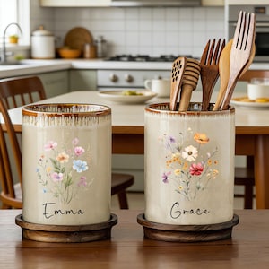 May include: Two ceramic utensil holders with floral designs. One is labeled "Emma" and the other "Grace". Both are beige with a brown rim and sit on wooden bases. The holder on the right contains wooden utensils.