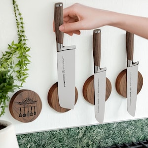 May include: Three kitchen knives with wooden handles and stainless steel blades are mounted on a white wall. The knives are held in place by round, wooden magnetic holders. The brand name "SCHMIDT BROS." is printed on the blades.