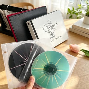 May include: A white binder with a black line drawing of a person playing guitar and the text Song Collection, accompanied by a black binder and two clear plastic sleeves containing a black disc with pink lines and a green disc with yellow lines.
