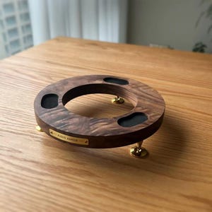 May include: A circular dark brown wood stand featuring three gold-toned metal feet and three black padded circular cutouts on the top surface. A small rectangular gold-toned metal plaque is attached to the front with the text AUDIO ARMOR.