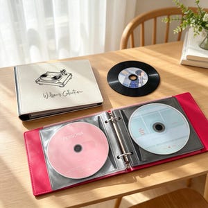 May include: A cream-colored CD binder with a black line drawing of a record player and the text "Willson's Collection" on the cover. An open bright pink binder displays two CDs, one pink and one light blue, alongside a loose black vinyl record.