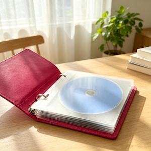 May include: A bright pink ring binder storage case featuring a textured cover and metal rings. Inside, the binder holds multiple clear plastic sleeves, each containing a single light blue disc. The binder is open, displaying the internal storage pages.