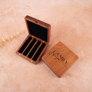 May include: A square wooden box with a hinged lid, featuring four rectangular slots lined with black velvet. The lid is engraved with the name Emily Johnson. One box is shown open, while the other is closed, displaying the wood grain finish.