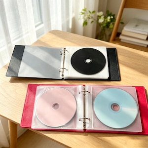 May include: Two open ring binder cases for discs, one black and one red, each featuring metal rings and clear plastic sleeves. The black binder holds a solid black disc, while the red binder contains one light pink disc and one light blue disc.