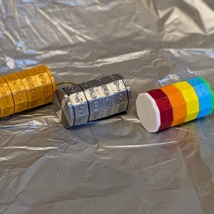 May include: Three different combination lock keychains. The first is gold with numbers 0-4, the second is silver with numbers 4-9, and the third is rainbow with numbers 0-9.