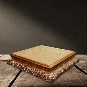 May include: A gold cake stand with an ornate, detailed design and a glittery surface. The stand is on a wooden surface.