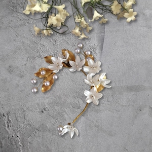 Könnte beinhalten: Ein goldener Blumenhaarschmuck mit weißen Blüten und Perlenakzenten. Der Haarschmuck ist halbmondförmig und perfekt für eine Hochzeit oder einen besonderen Anlass.