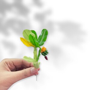 May include: A small, handmade clay sculpture of a banana plant with green leaves, yellow and orange bananas, and a red stem.