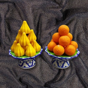 Pode incluir: Dois suportes de cerâmica decorativos com designs azuis e brancos. Um suporte contém uma pirâmide de sobremesas indianas amarelas e doces. O outro suporte contém uma pirâmide de frutas cítricas laranja.