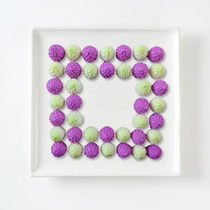 May include: A white square plate with a border of purple and green frosted cookies arranged in a square shape.