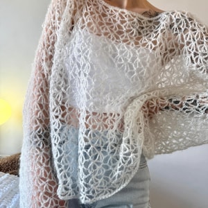 Puede incluir: Una parte superior blanca de manga larga y tejido abierto. La parte superior tiene un delicado patrón floral. La prenda es ligera y parece estar hecha de un material suave, posiblemente difuso. La parte superior se combina con vaqueros azules claros.