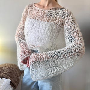 May include: A white, open-weave crochet top with long, flared sleeves. The top is worn over a white tank top and paired with light blue jeans. The crochet design creates a delicate, airy look.