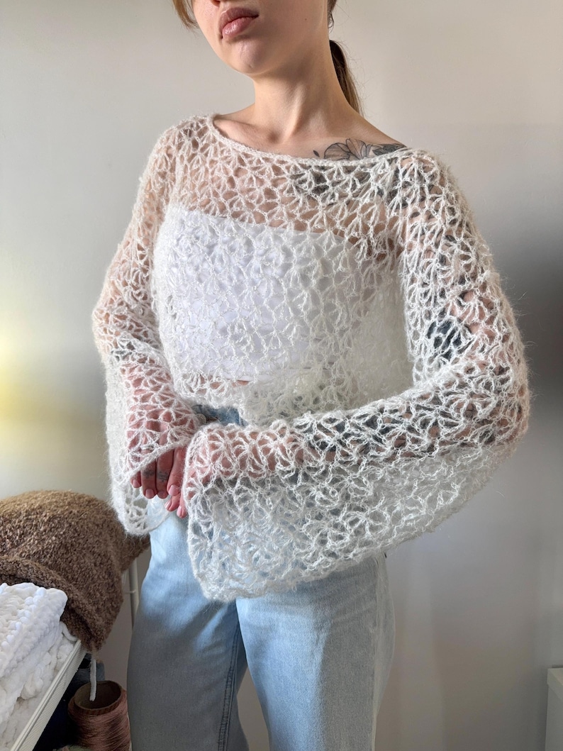 May include: A white, open-weave crochet top with long, flared sleeves. The top is worn over a white tank top and paired with light blue jeans. The crochet design creates a delicate, airy look.