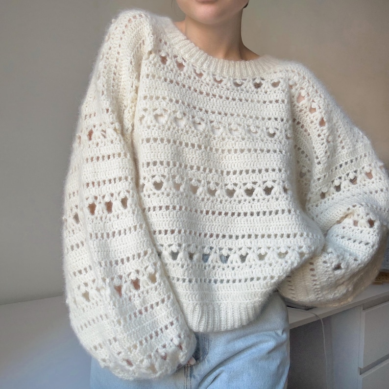 May include: A white crocheted sweater with a loose fit and a delicate openwork pattern. The sweater has long sleeves and a round neckline.