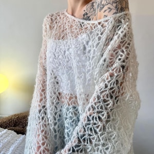 May include: A sheer, open-weave, off-white sweater with a round neckline and long sleeves. The sweater's delicate pattern creates a lacy effect, offering a glimpse of the garment worn underneath. This lightweight top is ideal for layering.