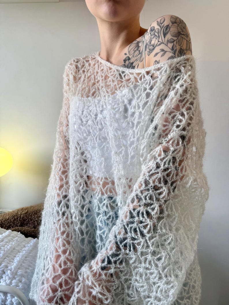 May include: A sheer, open-weave, off-white sweater with a round neckline and long sleeves. The sweater's delicate pattern creates a lacy effect, offering a glimpse of the garment worn underneath. This lightweight top is ideal for layering.